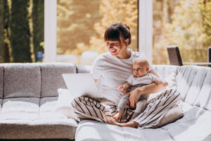 Mama pracująca na laptopie w domu, trzymając na kolanach niemowlę